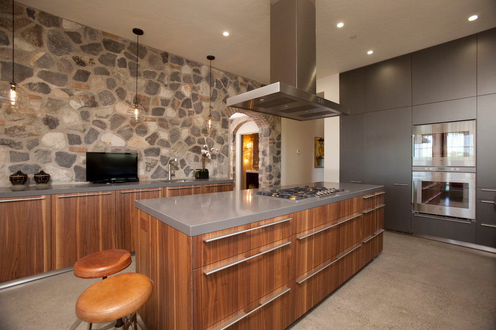 This is an example of a large industrial u-shaped enclosed kitchen in San Francisco with a submerged sink, flat-panel cabinets, medium wood cabinets, composite countertops, grey splashback, stone tiled splashback, stainless steel appliances, cement flooring and an island.