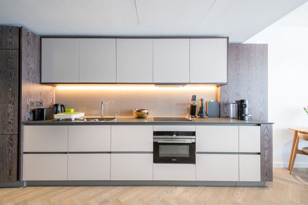 Example of a trendy single-wall light wood floor and beige floor kitchen design in London with an undermount sink, flat-panel cabinets, white cabinets, black appliances and gray countertops