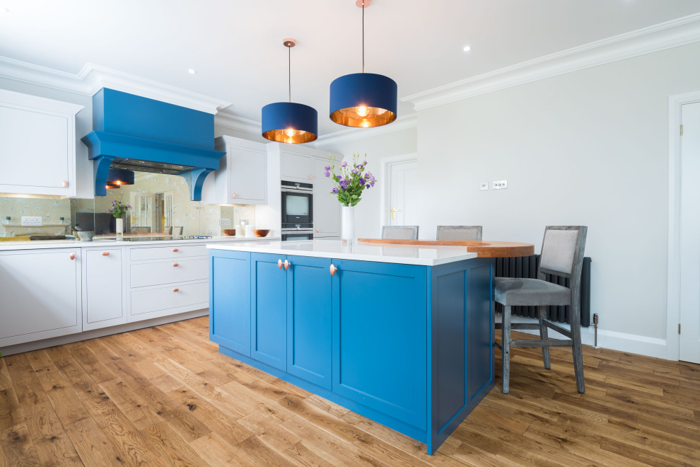 Classic l-shaped kitchen in London with shaker cabinets, blue cabinets, mirror splashback, black appliances, medium hardwood flooring, an island, brown floors and white worktops.