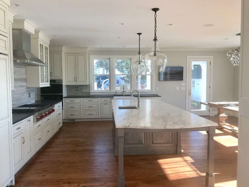 This is an example of a traditional kitchen/diner in Bridgeport with raised-panel cabinets, white cabinets, quartz worktops, white splashback, slate splashback, stainless steel appliances, medium hardwood flooring, an island and grey worktops.