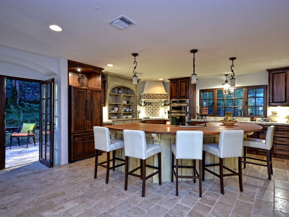 Large mountain style u-shaped travertine floor and beige floor eat-in kitchen photo in Austin with an island, raised-panel cabinets, dark wood cabinets, tile countertops, beige backsplash, ceramic backsplash, paneled appliances and a farmhouse sink