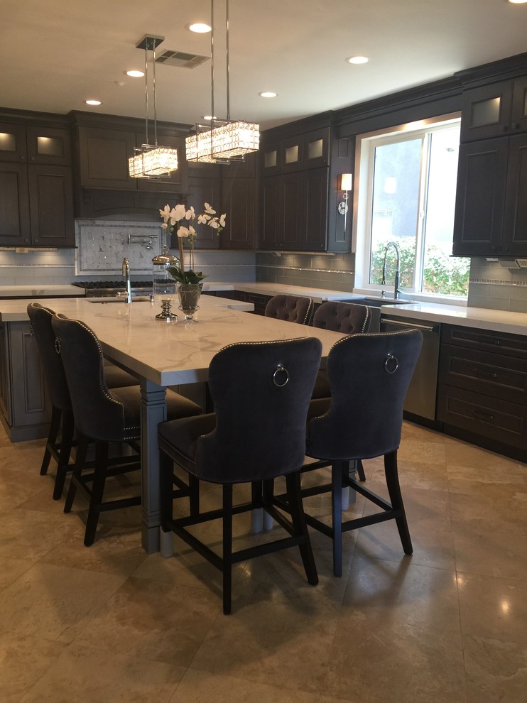 Mid-sized transitional l-shaped travertine floor and beige floor enclosed kitchen photo in Orange County with a farmhouse sink, beaded inset cabinets, brown cabinets, marble countertops, gray backsplash, glass tile backsplash, stainless steel appliances and an island