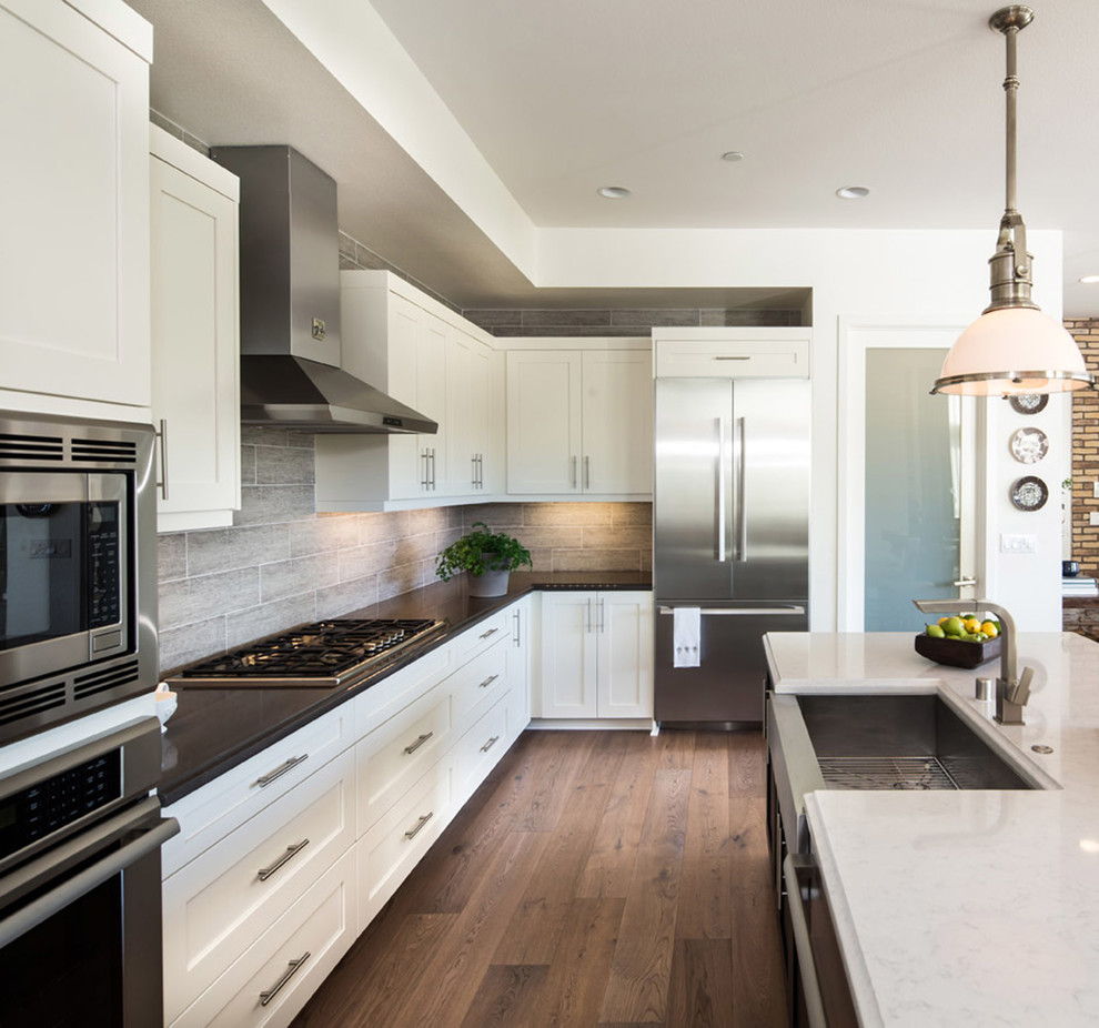 Example of a mid-sized cottage l-shaped medium tone wood floor eat-in kitchen design in Los Angeles with a farmhouse sink, shaker cabinets, white cabinets, quartz countertops, gray backsplash, porcelain backsplash, white appliances and an island