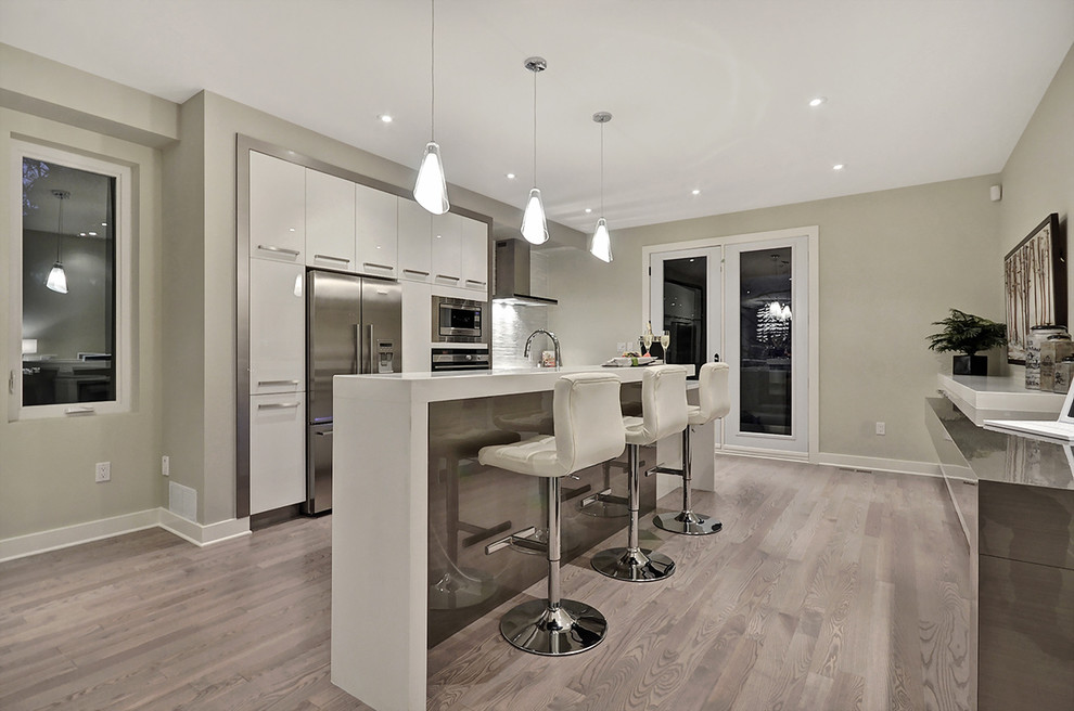 Example of a mid-sized trendy single-wall brown floor and laminate floor open concept kitchen design in Ottawa with flat-panel cabinets, white cabinets, solid surface countertops, stainless steel appliances, an island, an undermount sink, gray backsplash, matchstick tile backsplash and white countertops