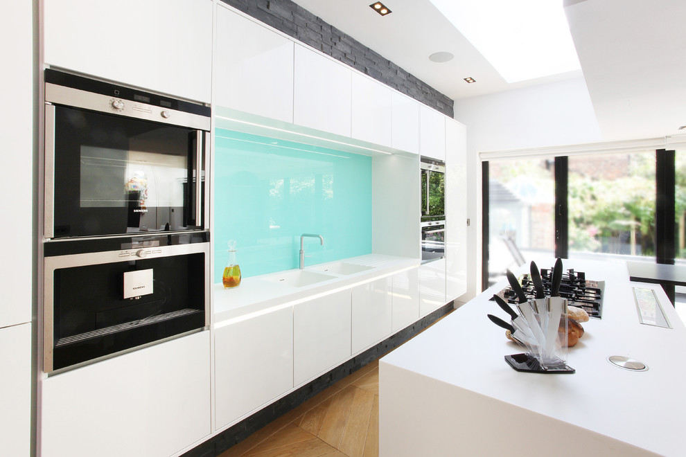 Example of a mid-sized trendy l-shaped light wood floor open concept kitchen design in Manchester with a drop-in sink, flat-panel cabinets, white cabinets and an island
