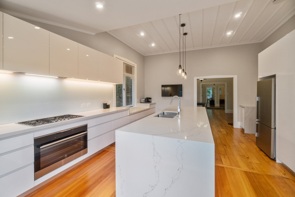 This is an example of an expansive modern single-wall kitchen/diner in Auckland with a double-bowl sink, white cabinets, composite countertops, white splashback, glass sheet splashback, stainless steel appliances, medium hardwood flooring, an island, brown floors, white worktops and flat-panel cabinets.