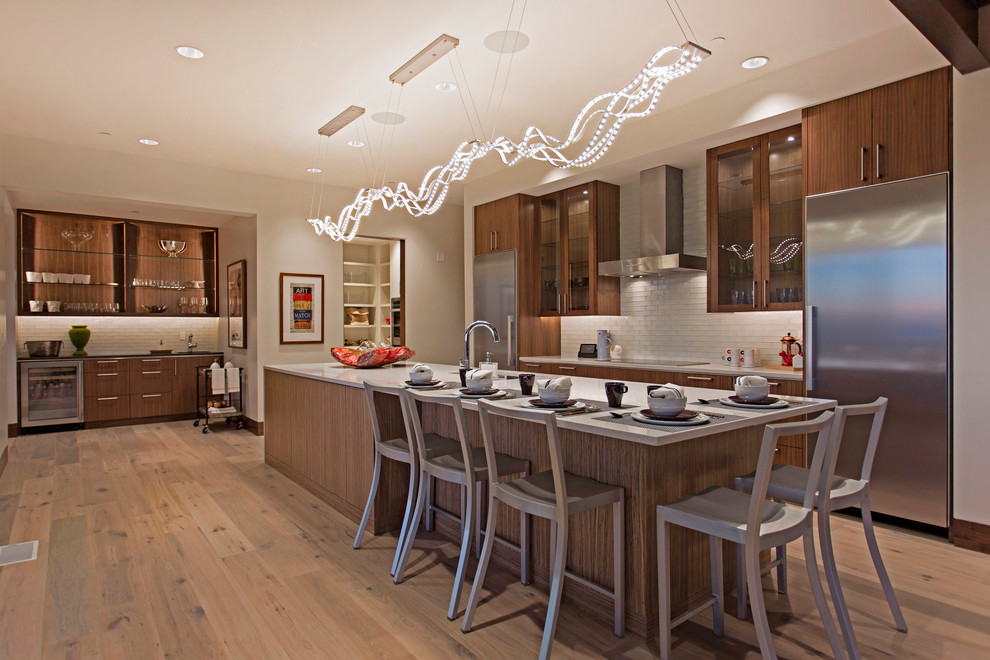 Large contemporary galley open plan kitchen in Salt Lake City with a submerged sink, flat-panel cabinets, medium wood cabinets, engineered stone countertops, white splashback, glass tiled splashback, light hardwood flooring, an island and stainless steel appliances.