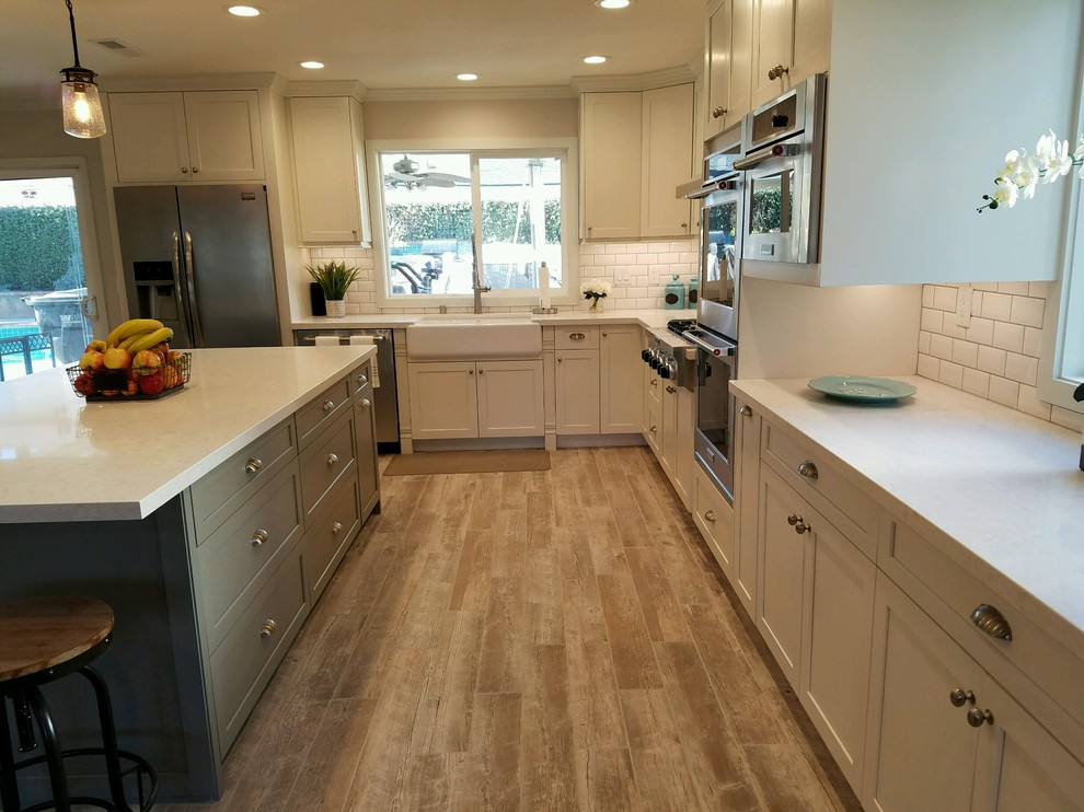 This is an example of a large traditional l-shaped open plan kitchen in Los Angeles with a belfast sink, shaker cabinets, white cabinets, engineered stone countertops, white splashback, metro tiled splashback, stainless steel appliances, light hardwood flooring, an island and brown floors.