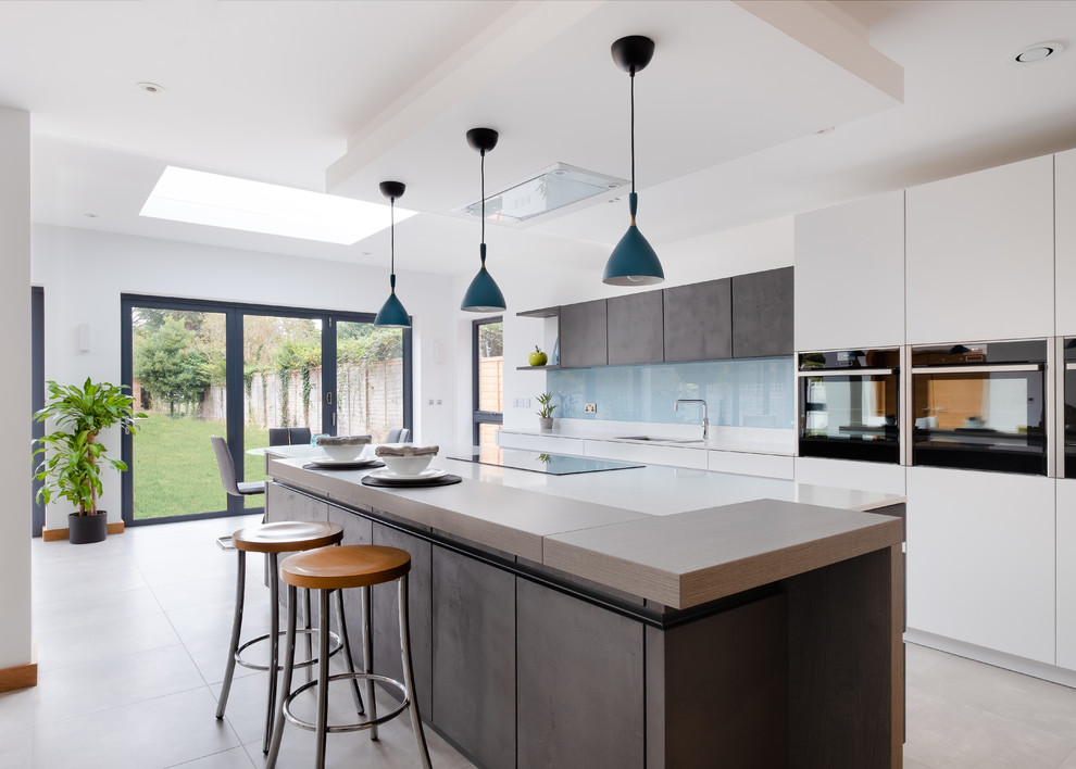 Large classic galley open plan kitchen in Gloucestershire with flat-panel cabinets, quartz worktops, blue splashback, glass sheet splashback, black appliances, porcelain flooring, an island, grey floors, white worktops, a submerged sink and white cabinets.