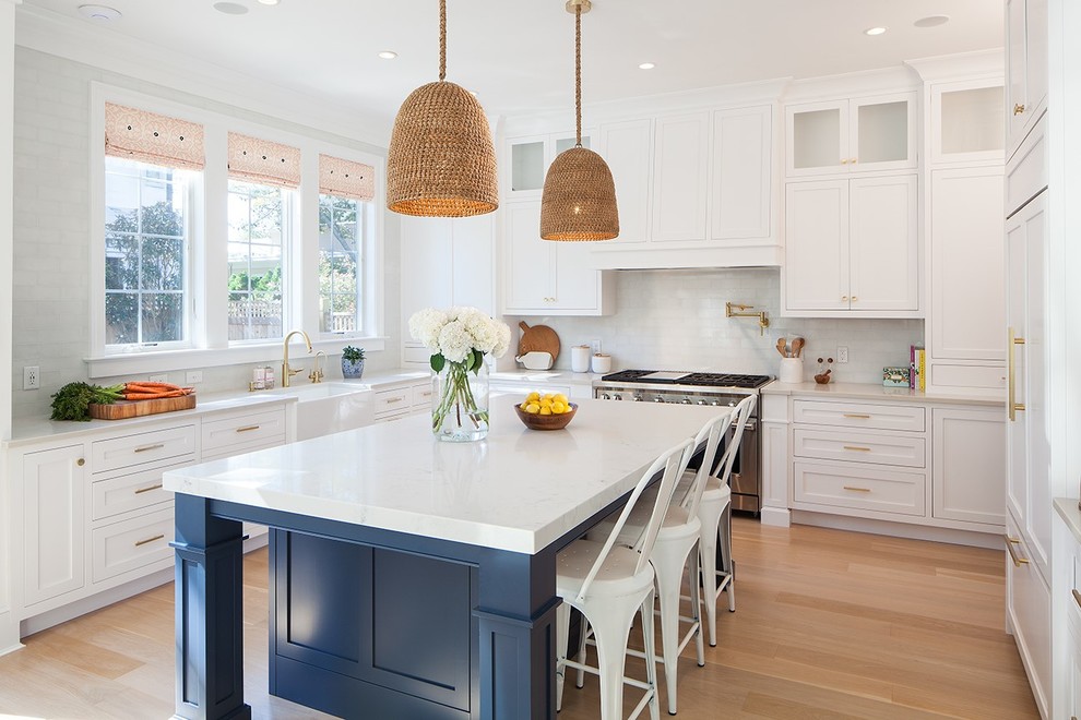 Inspiration for a coastal l-shaped kitchen in New York with a belfast sink, shaker cabinets, white cabinets, white splashback, stainless steel appliances, light hardwood flooring and an island.