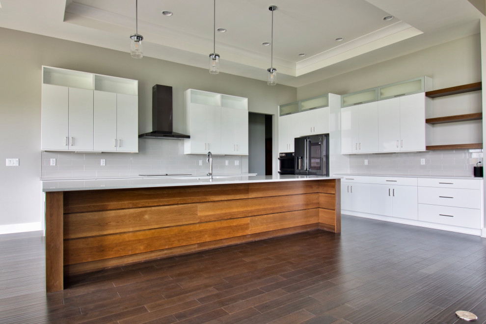 Large modern l-shaped open plan kitchen in Milwaukee with a belfast sink, flat-panel cabinets, white cabinets, engineered stone countertops, grey splashback, metro tiled splashback, black appliances, dark hardwood flooring, an island, brown floors, white worktops and a drop ceiling.