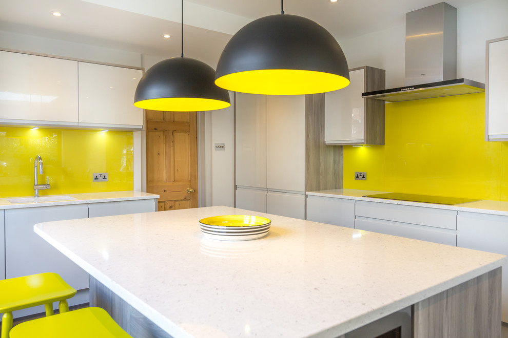 Photo of a contemporary kitchen/diner in London with a built-in sink, flat-panel cabinets, grey cabinets, composite countertops, yellow splashback, glass sheet splashback, stainless steel appliances, porcelain flooring and an island.