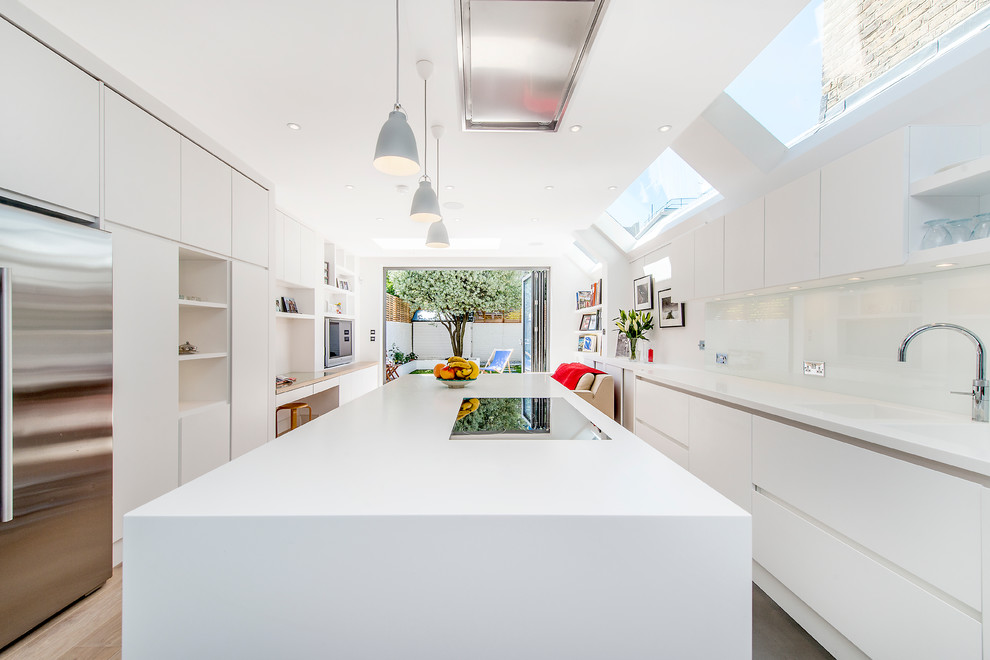 Large contemporary galley open plan kitchen in London with a built-in sink, flat-panel cabinets, white cabinets, composite countertops, white splashback, glass sheet splashback, stainless steel appliances, medium hardwood flooring and an island.