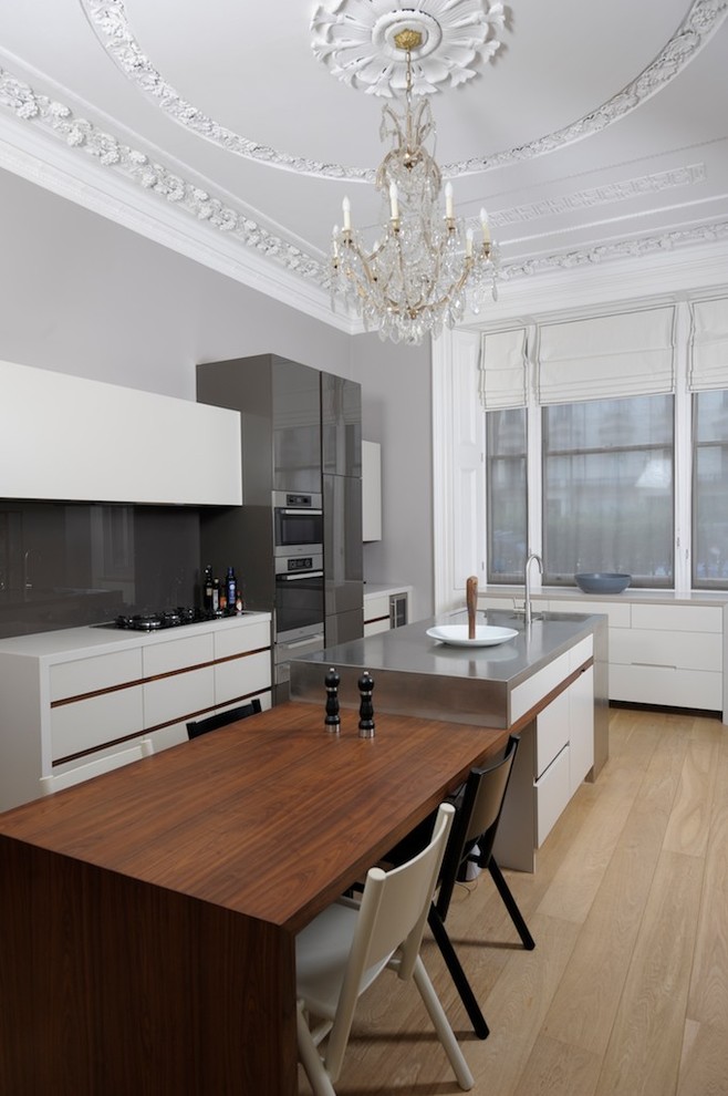 Large contemporary galley kitchen/diner in London with an integrated sink, flat-panel cabinets, white cabinets, composite countertops, grey splashback, glass sheet splashback, stainless steel appliances, light hardwood flooring and an island.