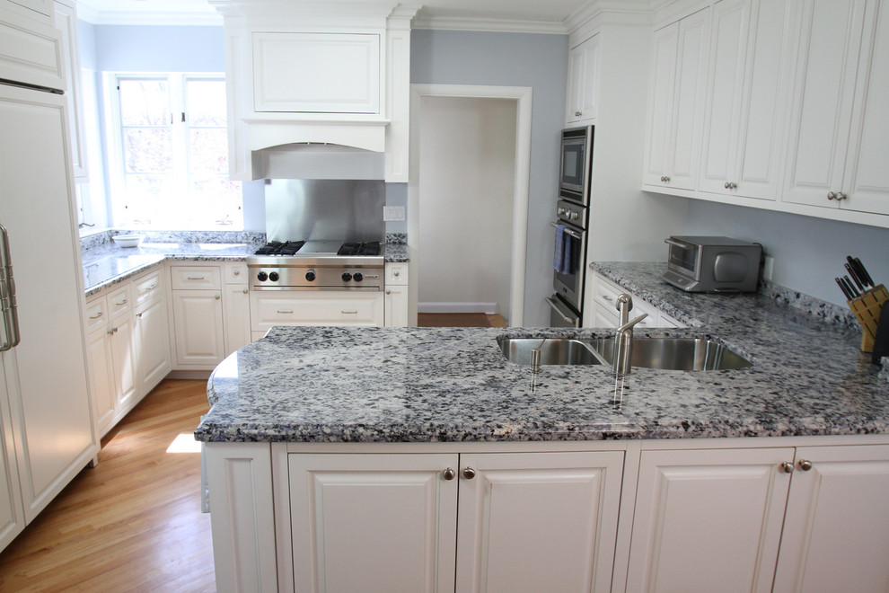 Klassisk inredning av ett stort kök, med en undermonterad diskho, luckor med upphöjd panel, vita skåp, granitbänkskiva, integrerade vitvaror, ljust trägolv och en halv köksö