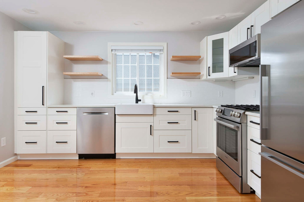 ボストンにあるラグジュアリーな中くらいなモダンスタイルのおしゃれな独立型キッチン (エプロンフロントシンク、シェーカースタイル扉のキャビネット、白いキャビネット、クオーツストーンカウンター、白いキッチンパネル、セラミックタイルのキッチンパネル、シルバーの調理設備、淡色無垢フローリング、白いキッチンカウンター) の写真