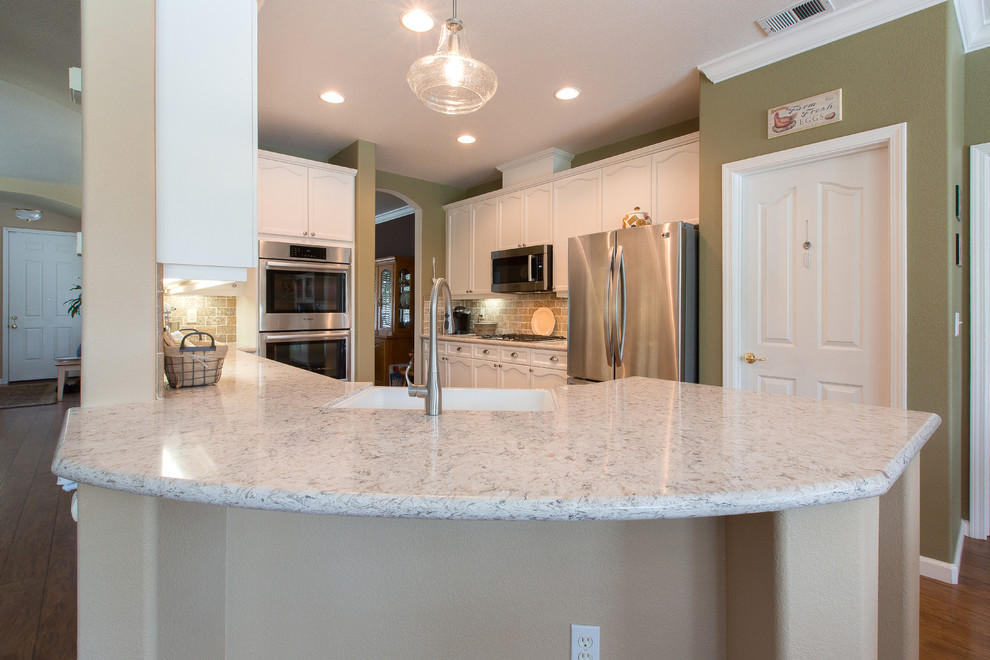 Cette image montre une cuisine minimaliste avec un évier de ferme, des portes de placard blanches, un plan de travail en quartz modifié, une crédence beige, un électroménager en acier inoxydable et une péninsule.