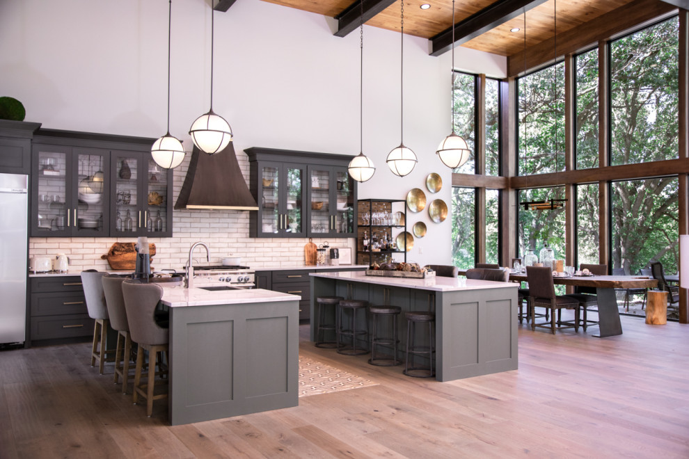 Example of a large transitional u-shaped medium tone wood floor and brown floor open concept kitchen design in Other with a farmhouse sink, shaker cabinets, black cabinets, quartzite countertops, white backsplash, porcelain backsplash, stainless steel appliances, two islands and white countertops