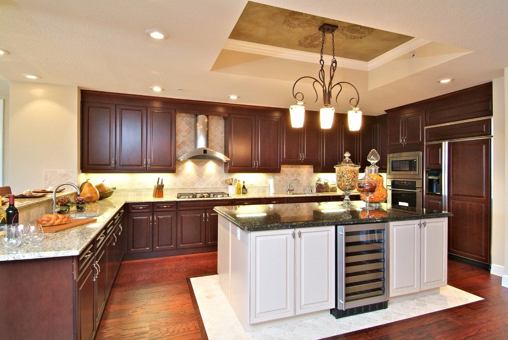 Example of a mid-sized island style l-shaped medium tone wood floor eat-in kitchen design in Orlando with an undermount sink, raised-panel cabinets, dark wood cabinets, granite countertops, beige backsplash, stone tile backsplash, stainless steel appliances and an island
