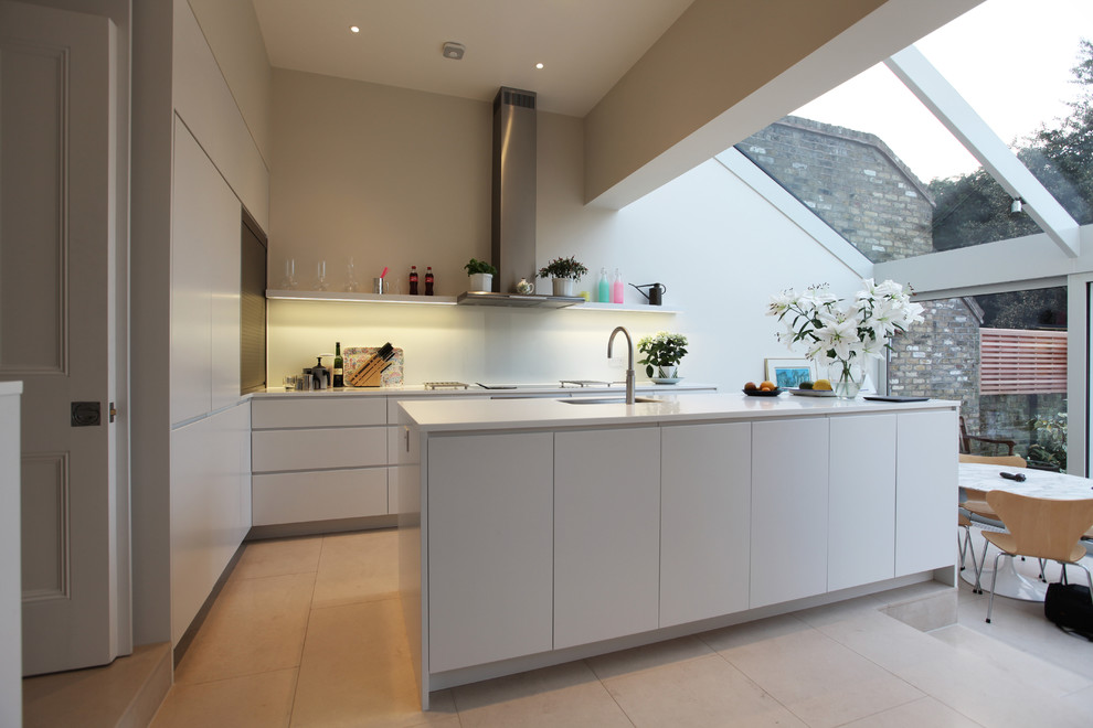 Foto på ett kök, med en enkel diskho, släta luckor, vita skåp, glaspanel som stänkskydd, kalkstensgolv och en köksö