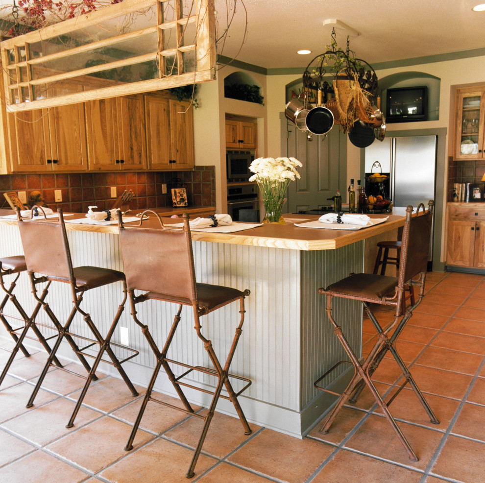 Bild på ett rustikt kök och matrum, med skåp i mellenmörkt trä, träbänkskiva, rött stänkskydd, stänkskydd i terrakottakakel, rostfria vitvaror, klinkergolv i terrakotta och en halv köksö