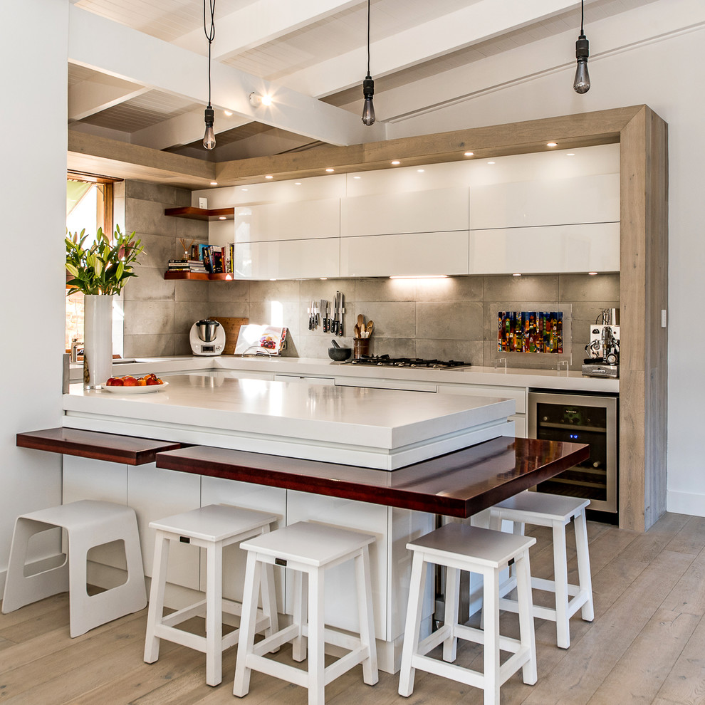 Foto på ett funkis vit u-kök, med släta luckor, vita skåp, grått stänkskydd, rostfria vitvaror, ljust trägolv, en halv köksö och beiget golv