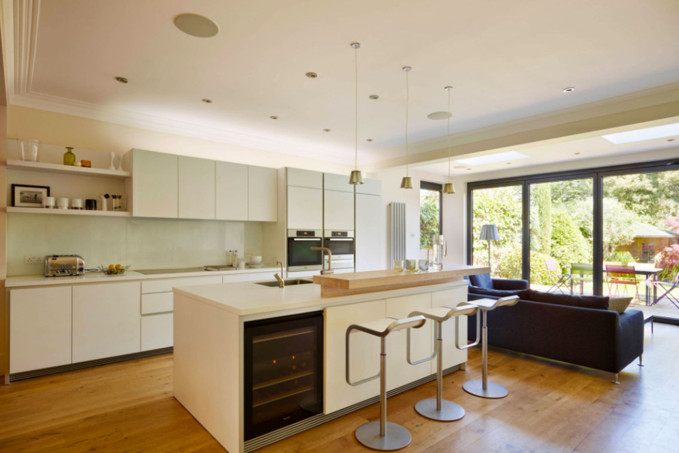Medium sized contemporary l-shaped open plan kitchen in DC Metro with a submerged sink, flat-panel cabinets, white cabinets, engineered stone countertops, white splashback, stainless steel appliances, light hardwood flooring, an island, beige floors, glass sheet splashback and white worktops.