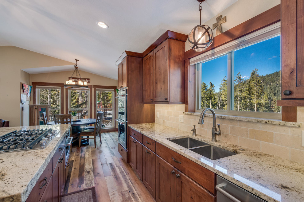 This is an example of a large rustic l-shaped enclosed kitchen in Denver with a submerged sink, shaker cabinets, medium wood cabinets, granite worktops, beige splashback, stone tiled splashback, stainless steel appliances, medium hardwood flooring, an island and brown floors.