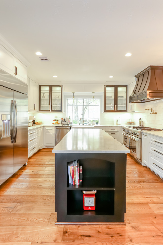 Inspiration for a large transitional u-shaped medium tone wood floor and brown floor enclosed kitchen remodel in Houston with a farmhouse sink, raised-panel cabinets, white cabinets, granite countertops, white backsplash, glass tile backsplash, stainless steel appliances, an island and white countertops