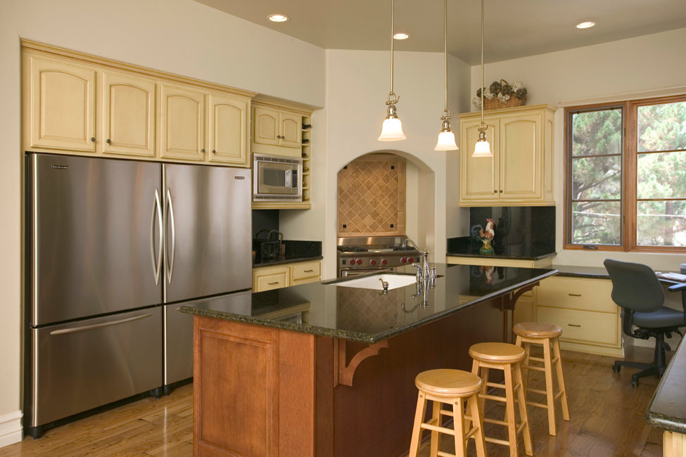 Inredning av ett lantligt kök, med en undermonterad diskho, luckor med upphöjd panel, gula skåp, granitbänkskiva, svart stänkskydd, stänkskydd i sten, rostfria vitvaror, mellanmörkt trägolv och en köksö