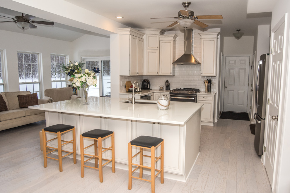 Inspiration for a mid-sized timeless l-shaped light wood floor eat-in kitchen remodel in New York with an undermount sink, recessed-panel cabinets, white cabinets, quartz countertops, white backsplash, ceramic backsplash, stainless steel appliances and an island
