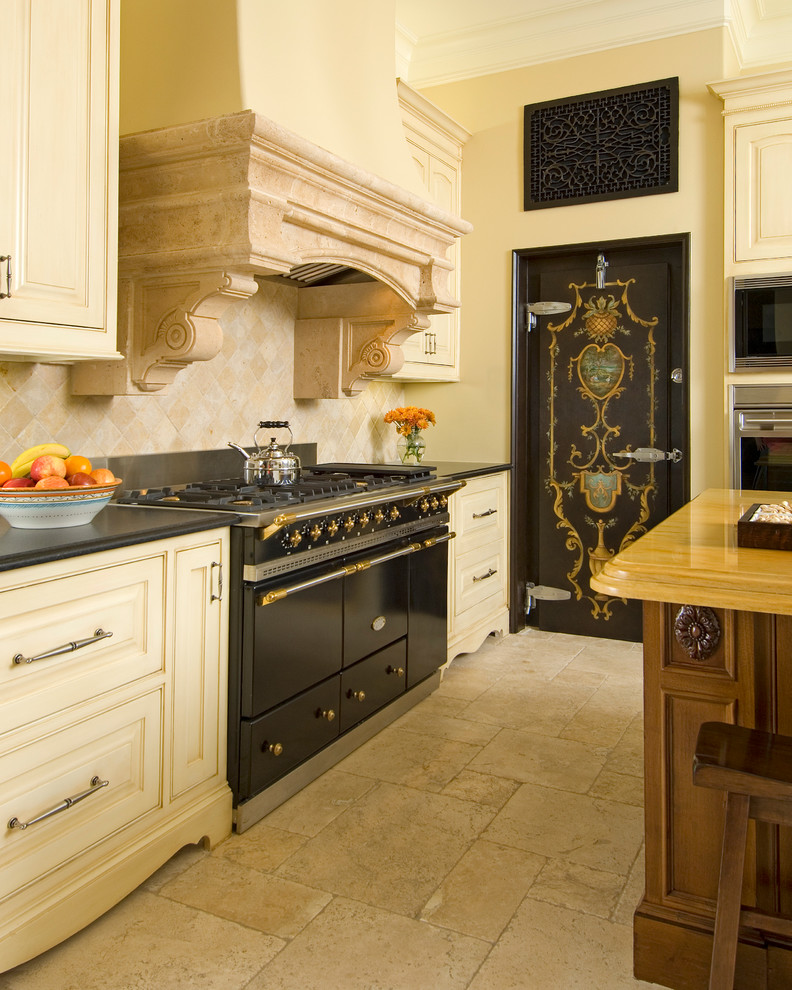 Photo of a mediterranean kitchen in Dallas with raised-panel cabinets, beige cabinets, beige splashback, black appliances, an island and beige floors.