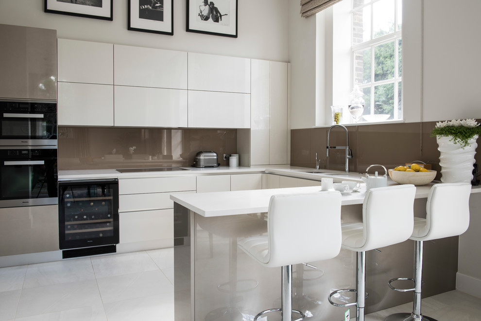 Contemporary u-shaped kitchen/diner in Hertfordshire with a submerged sink, flat-panel cabinets, white cabinets, brown splashback, glass sheet splashback, black appliances, a breakfast bar, white floors and white worktops.