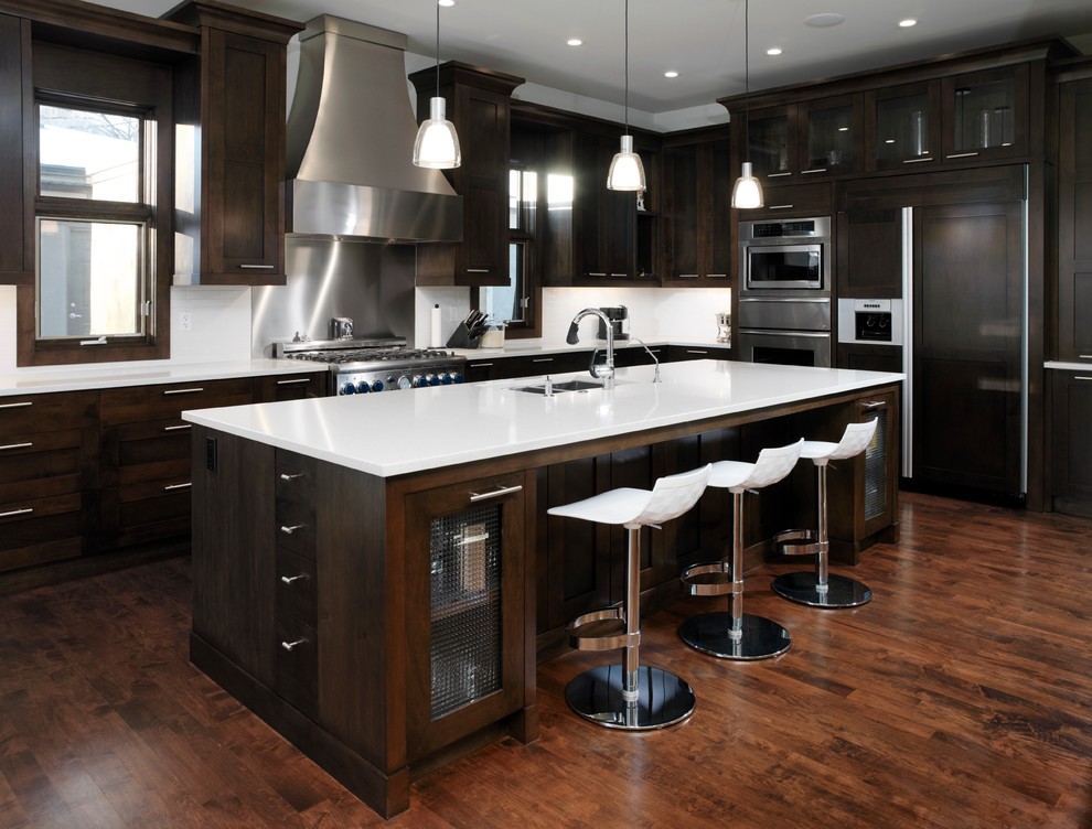 Modelo de cocina contemporánea grande con armarios estilo shaker, puertas de armario de madera en tonos medios, salpicadero blanco, electrodomésticos de acero inoxidable, suelo de madera en tonos medios y una isla