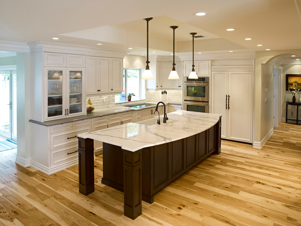 Large beach style l-shaped light wood floor eat-in kitchen photo in Miami with a farmhouse sink, raised-panel cabinets, white cabinets, marble countertops, white backsplash, subway tile backsplash and an island