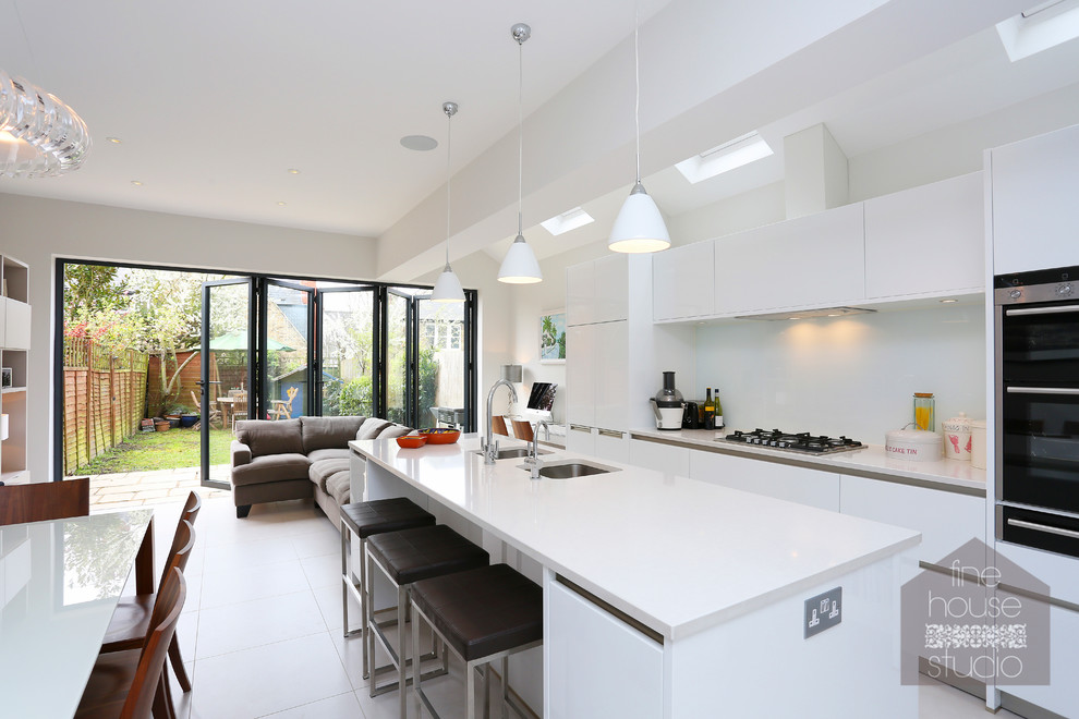Large contemporary kitchen/diner in London with a double-bowl sink, white cabinets, engineered stone countertops, glass sheet splashback, ceramic flooring and an island.