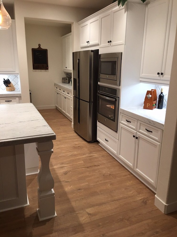 Photo of a medium sized classic l-shaped open plan kitchen in Los Angeles with a belfast sink, recessed-panel cabinets, white cabinets, engineered stone countertops, white splashback, ceramic splashback, stainless steel appliances, medium hardwood flooring, an island, brown floors and grey worktops.