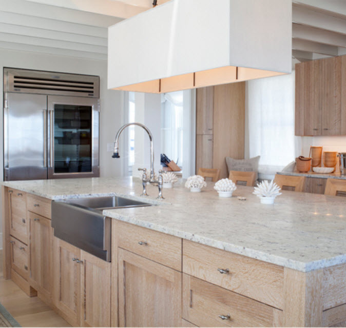 Large beach style enclosed kitchen in Chicago with a belfast sink, flat-panel cabinets, light wood cabinets, engineered stone countertops, white splashback, glass tiled splashback, stainless steel appliances, light hardwood flooring and an island.