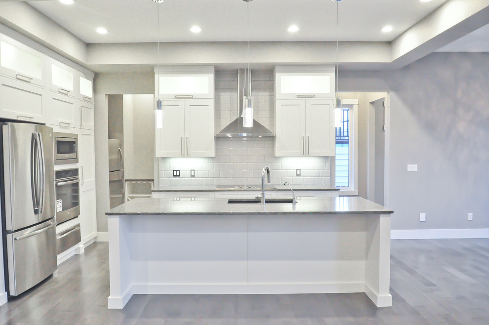 Bild på ett mellanstort funkis kök, med en nedsänkt diskho, luckor med glaspanel, vita skåp, granitbänkskiva, grått stänkskydd, stänkskydd i glaskakel, rostfria vitvaror, mellanmörkt trägolv, en köksö och grått golv