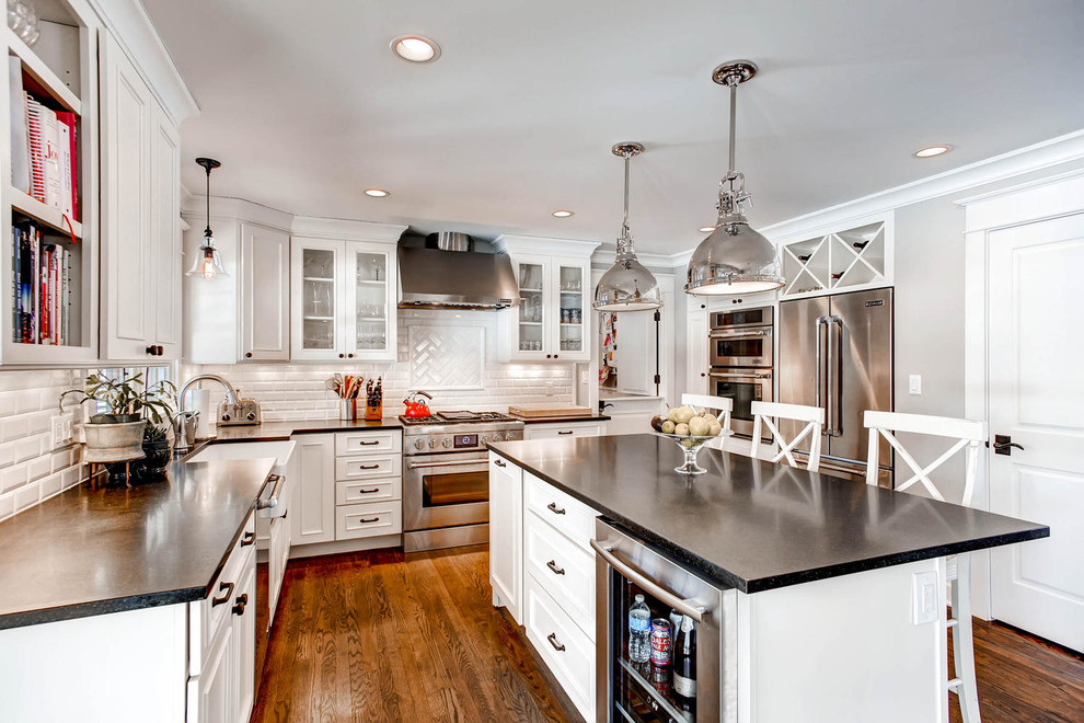 This is an example of a traditional u-shaped kitchen in Denver with a belfast sink, recessed-panel cabinets, white cabinets, white splashback, metro tiled splashback, stainless steel appliances, dark hardwood flooring and an island.