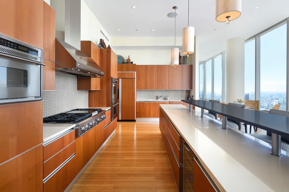 Modern inredning av ett l-kök, med en undermonterad diskho, släta luckor, skåp i mellenmörkt trä, grått stänkskydd, stänkskydd i glaskakel, rostfria vitvaror, mellanmörkt trägolv och en köksö