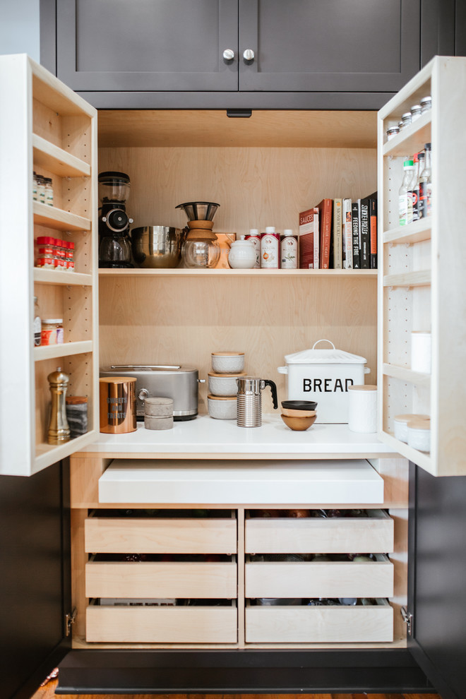 This is an example of a medium sized l-shaped kitchen pantry in Atlanta with a belfast sink, shaker cabinets, black cabinets, engineered stone countertops, white splashback, cement tile splashback, stainless steel appliances, medium hardwood flooring, an island, brown floors and white worktops.
