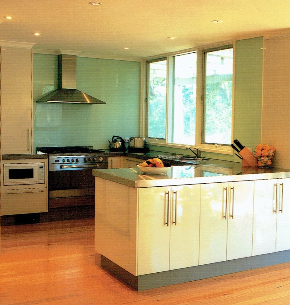 Photo of a medium sized contemporary u-shaped kitchen in Canberra - Queanbeyan with flat-panel cabinets, white cabinets, a submerged sink, stainless steel worktops, green splashback, glass tiled splashback, stainless steel appliances and medium hardwood flooring.