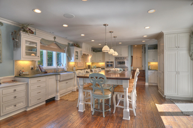 Example of a beach style l-shaped medium tone wood floor eat-in kitchen design in Orange County with a farmhouse sink, recessed-panel cabinets, white cabinets, granite countertops, beige backsplash, stone tile backsplash, stainless steel appliances and no island