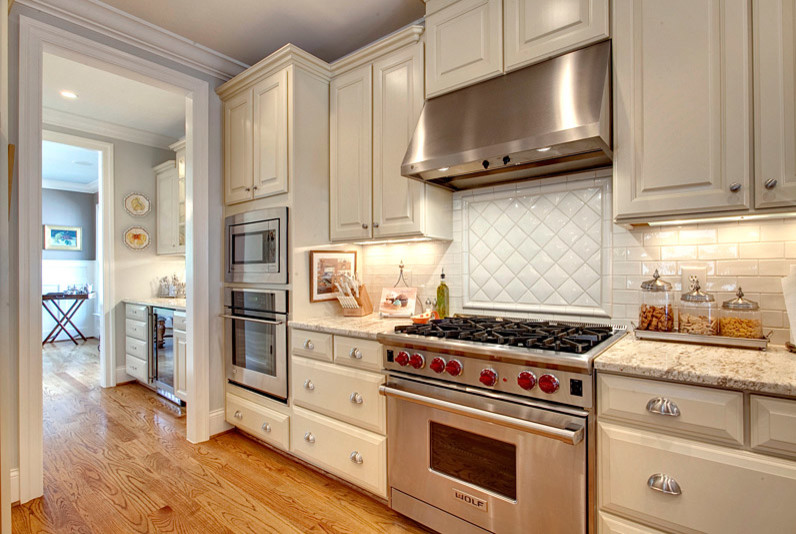 Imagen de cocina clásica con puertas de armario blancas, encimera de granito, salpicadero blanco, salpicadero de azulejos tipo metro, electrodomésticos de acero inoxidable y suelo de madera clara
