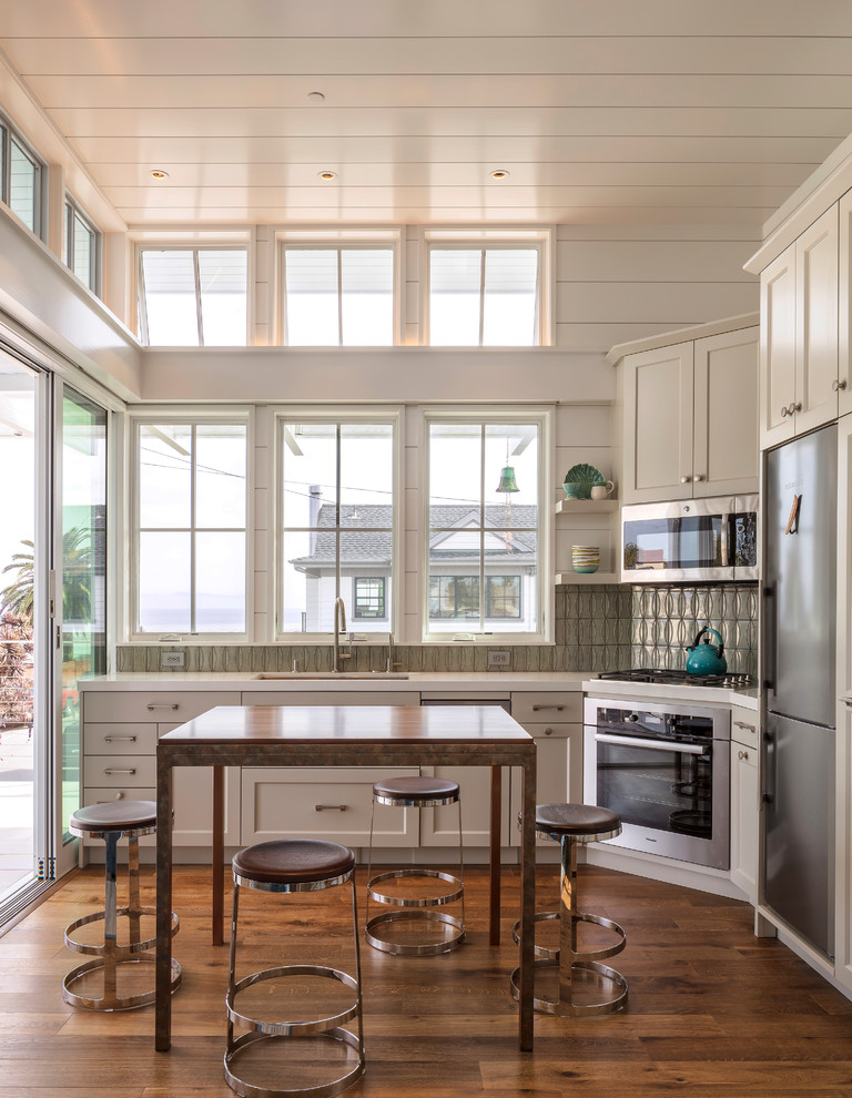 Coastal l-shaped kitchen/diner in San Luis Obispo with a submerged sink, shaker cabinets, white cabinets, grey splashback, stainless steel appliances, medium hardwood flooring, brown floors and white worktops.