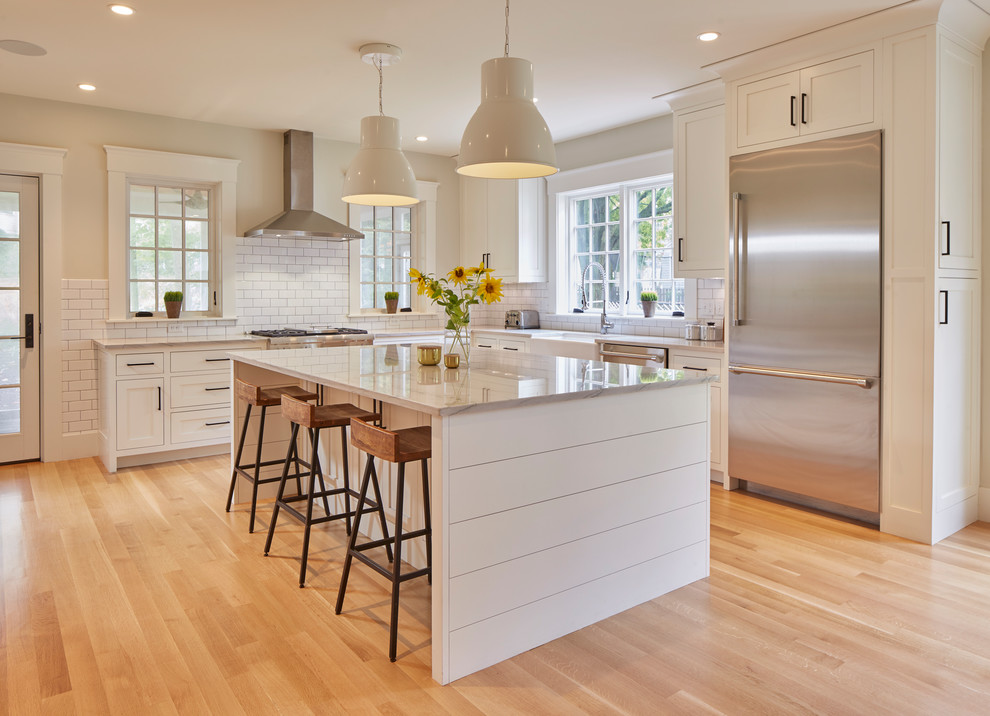 Design ideas for a traditional l-shaped kitchen in Boston with a belfast sink, shaker cabinets, white cabinets, white splashback, metro tiled splashback, stainless steel appliances, medium hardwood flooring, an island, brown floors and white worktops.