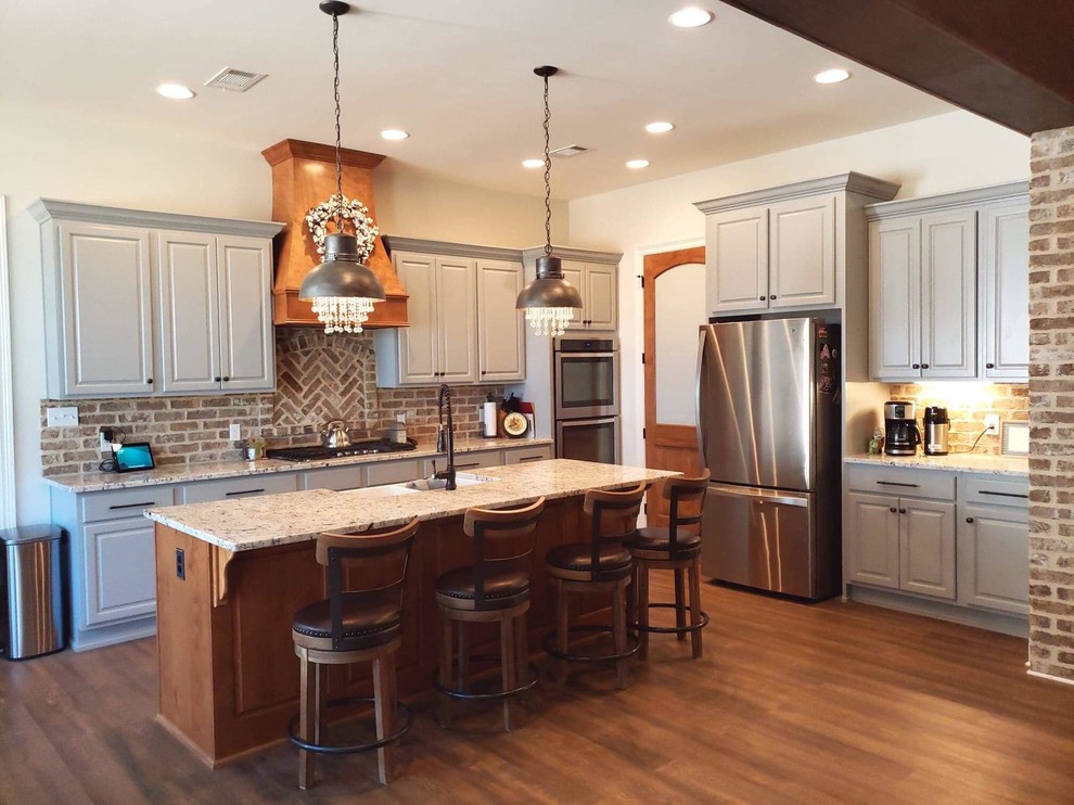 Photo of a medium sized farmhouse l-shaped open plan kitchen in New Orleans with a belfast sink, raised-panel cabinets, grey cabinets, brick splashback, stainless steel appliances and an island.