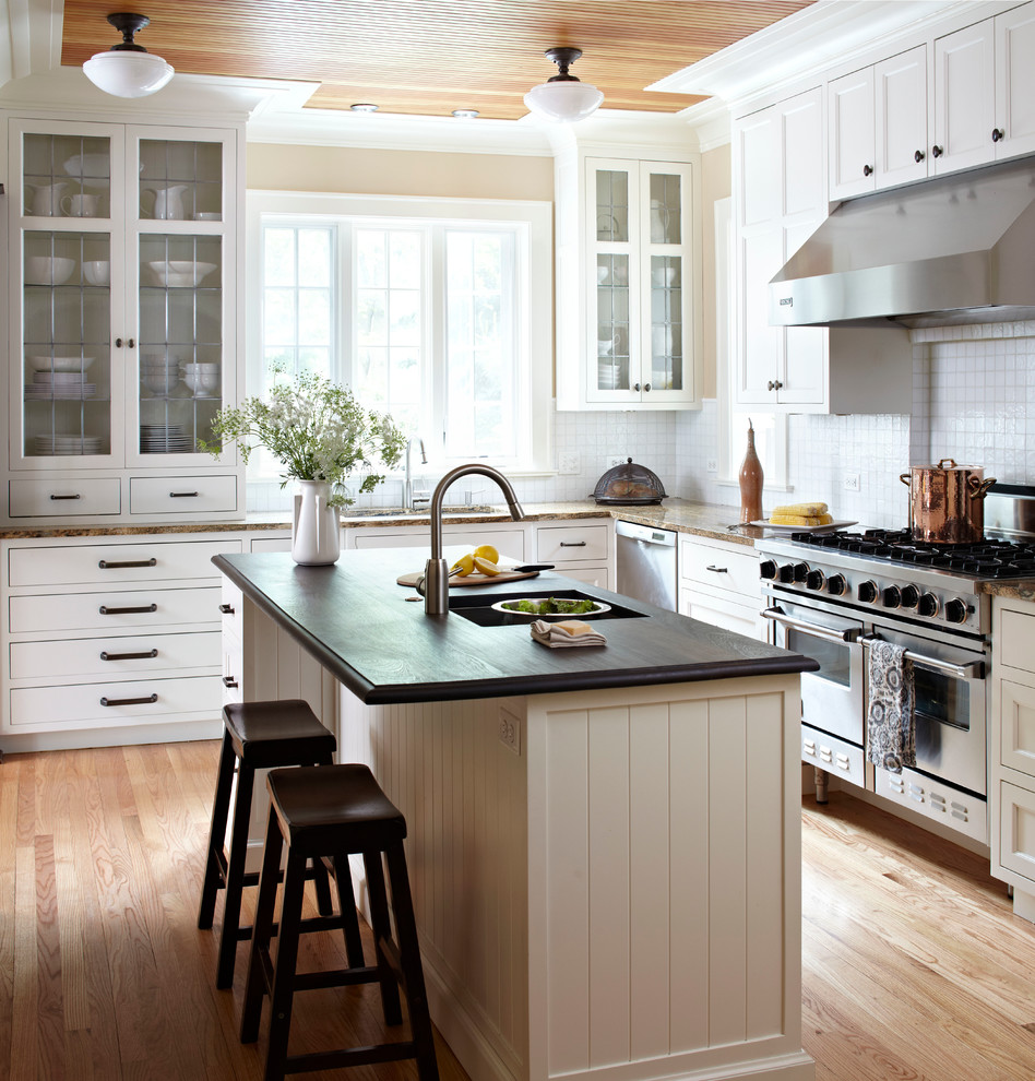 Cette image montre une cuisine américaine traditionnelle en L avec un évier encastré, un placard à porte vitrée, des portes de placard blanches, un plan de travail en bois, une crédence blanche et un sol en bois brun.
