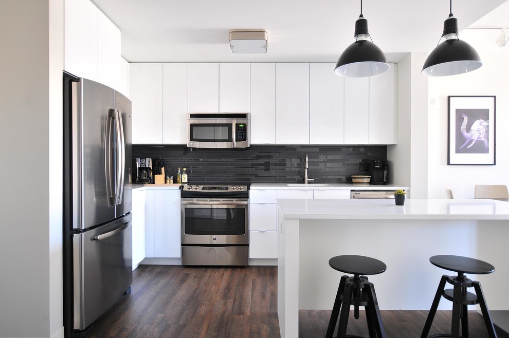 Exempel på ett modernt vit vitt l-kök, med släta luckor, vita skåp, svart stänkskydd, rostfria vitvaror, mörkt trägolv, en köksö och brunt golv